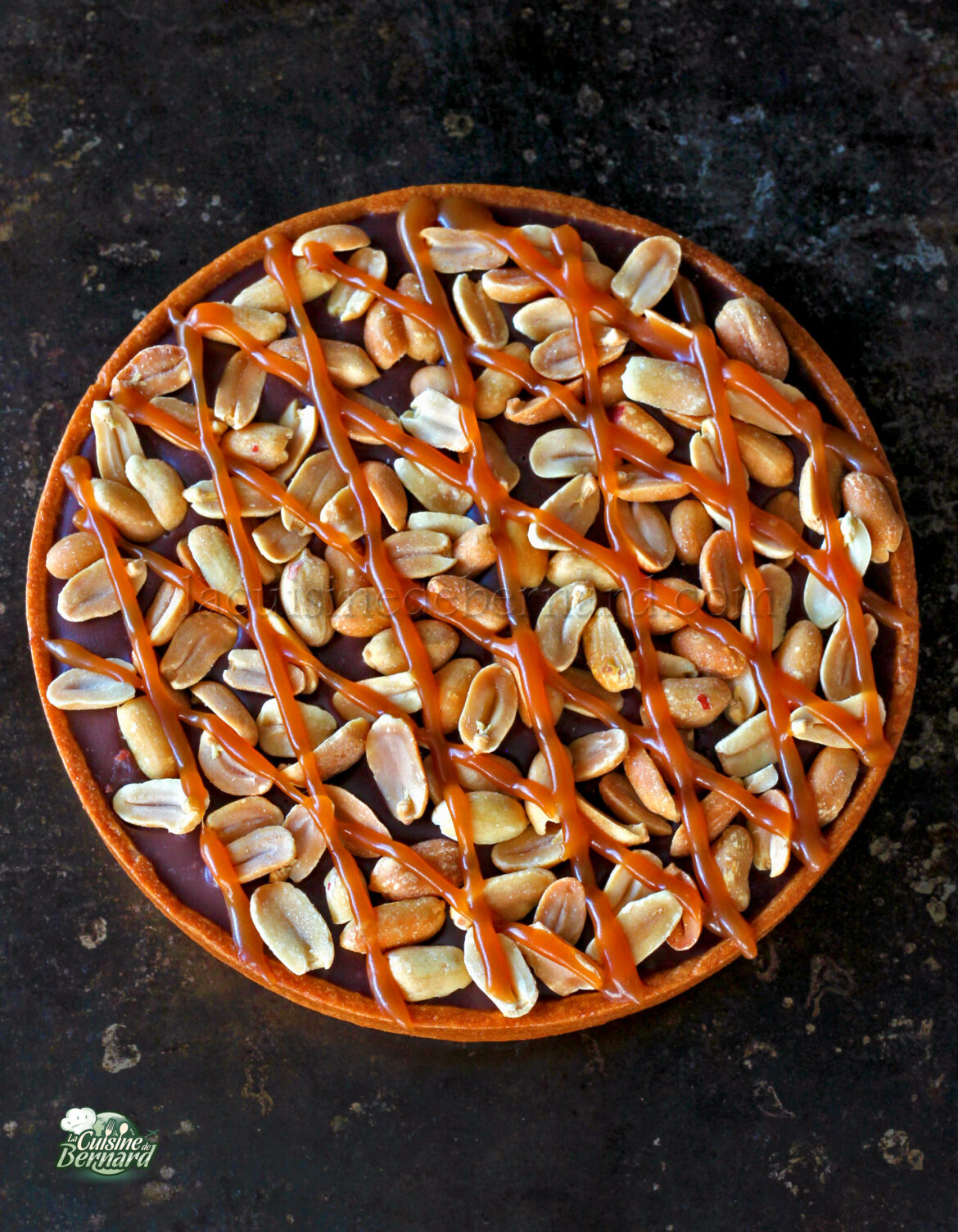 Tarte ronde garnie de cacahuètes et arrosée de caramel sur une surface foncée.