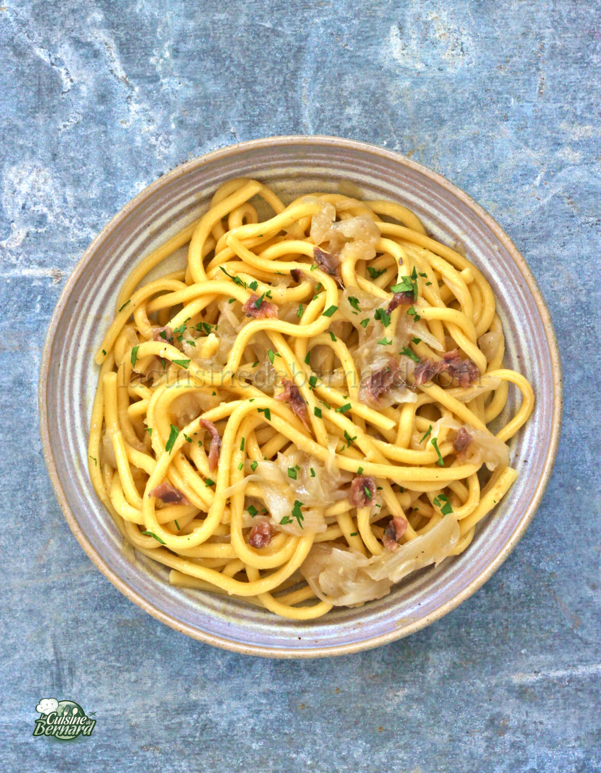 Un bol de pâtes Bigoli aux nouilles épaisses, garni d'herbes et mélangé à des oignons caramélisés et des petits morceaux de viande. Le plat est joliment présenté sur une surface grise texturée, subtilement rehaussée d'une touche de salsa pour une saveur plus intense.