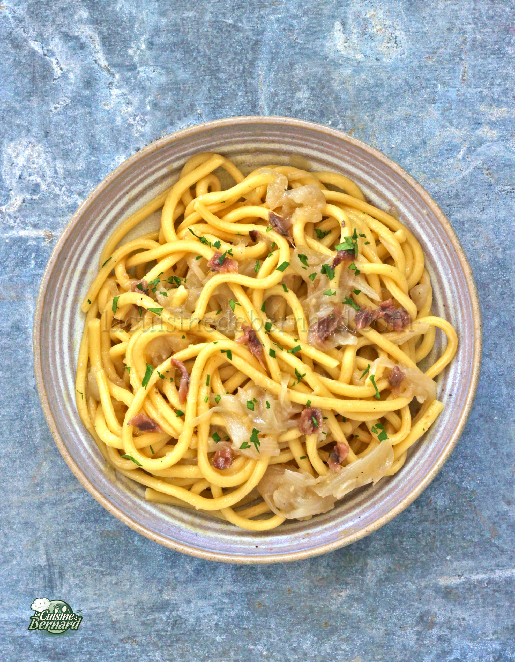 Un bol de pâtes Bigoli aux nouilles épaisses, garni d'herbes et mélangé à des oignons caramélisés et des petits morceaux de viande. Le plat est joliment présenté sur une surface grise texturée, subtilement rehaussée d'une touche de salsa pour une saveur plus intense.