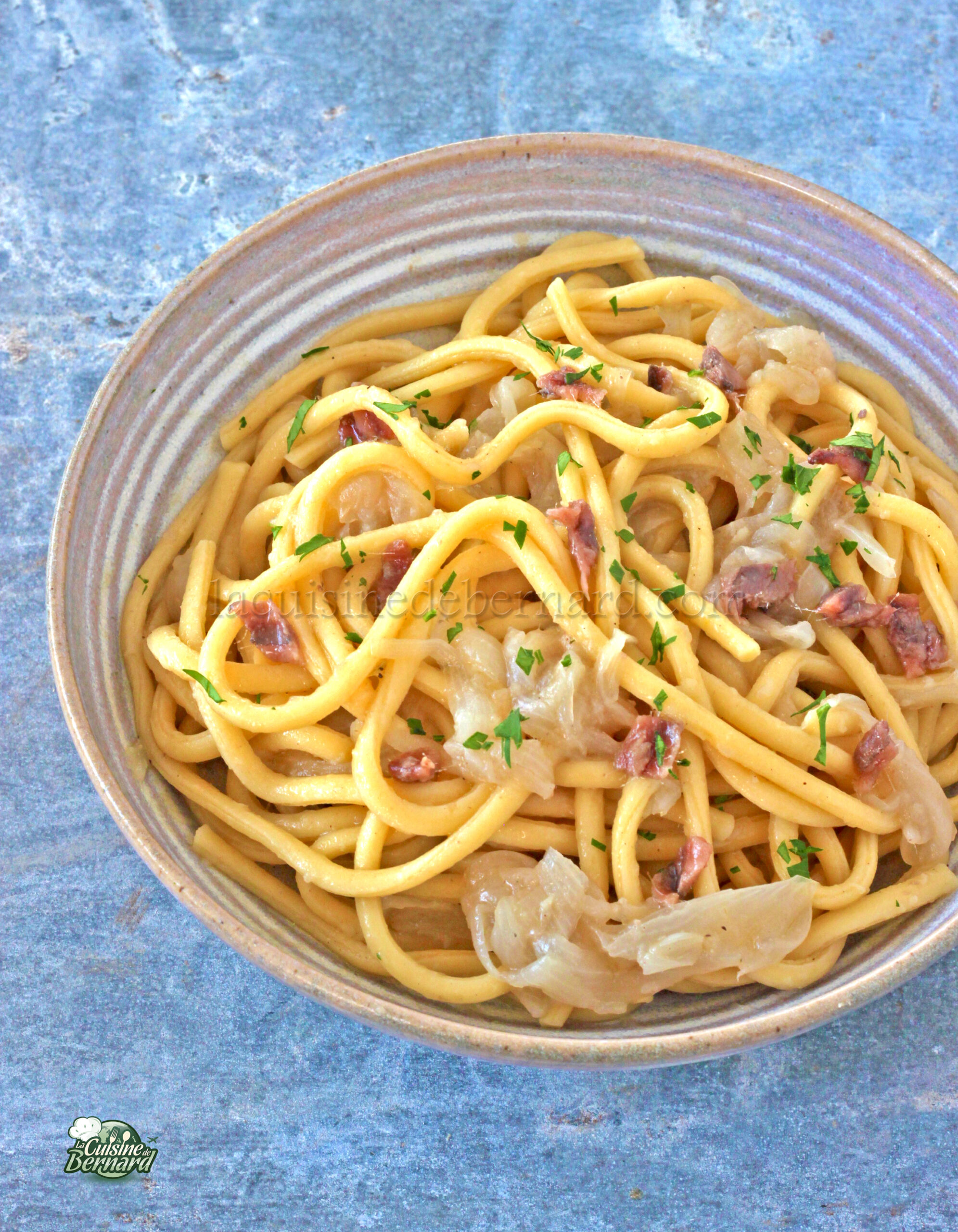 Un bol de pâtes italiennes garni d'oignons caramélisés, de petits morceaux de viande et de persil haché repose sur une surface bleu gris texturée, rappelant un bigoli traditionnel en salsa.
