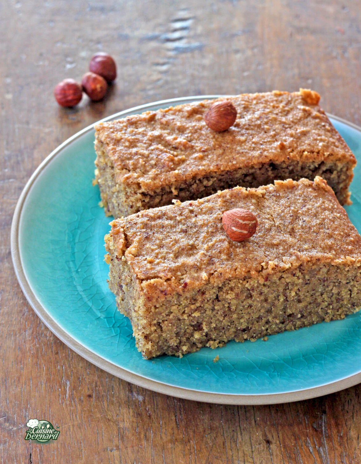 Deux tranches de gâteau financier brun noisette garnies de noisettes entières, élégamment servies sur une assiette turquoise posée sur une table en bois.