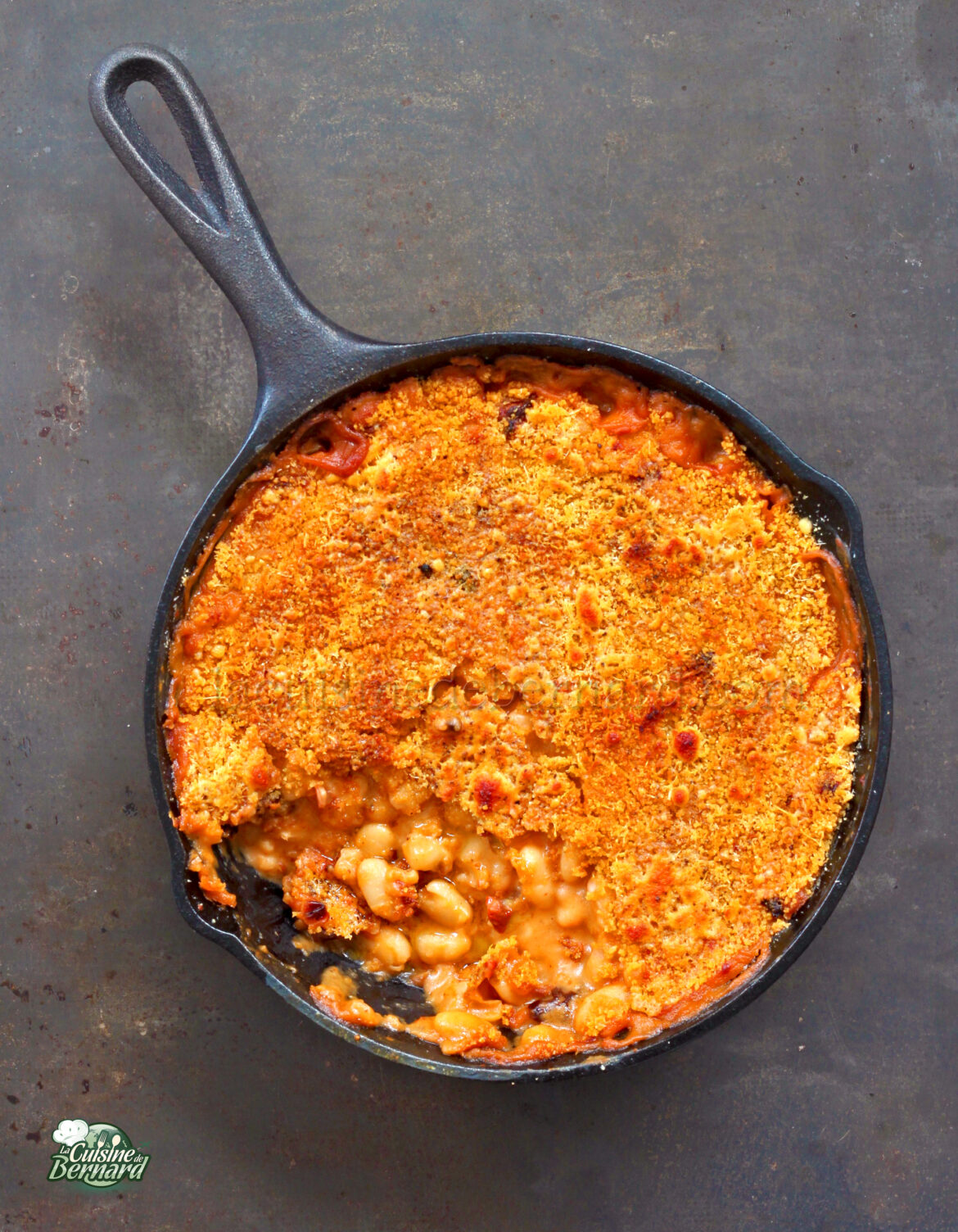 Une poêle en fonte remplie de haricots blancs à la sauce tomate, garnie d'une croûte de chapelure dorée croustillante et d'un soupçon de tomates séchées, avec une partie retirée pour révéler la garniture copieuse.