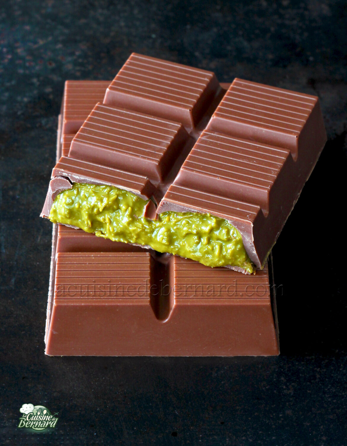 Une tablette de chocolat au lait Chocolat de Dubaï est cassée en deux, révélant une onctueuse crème à la pistache verte. Le chocolat repose élégamment sur une surface sombre.