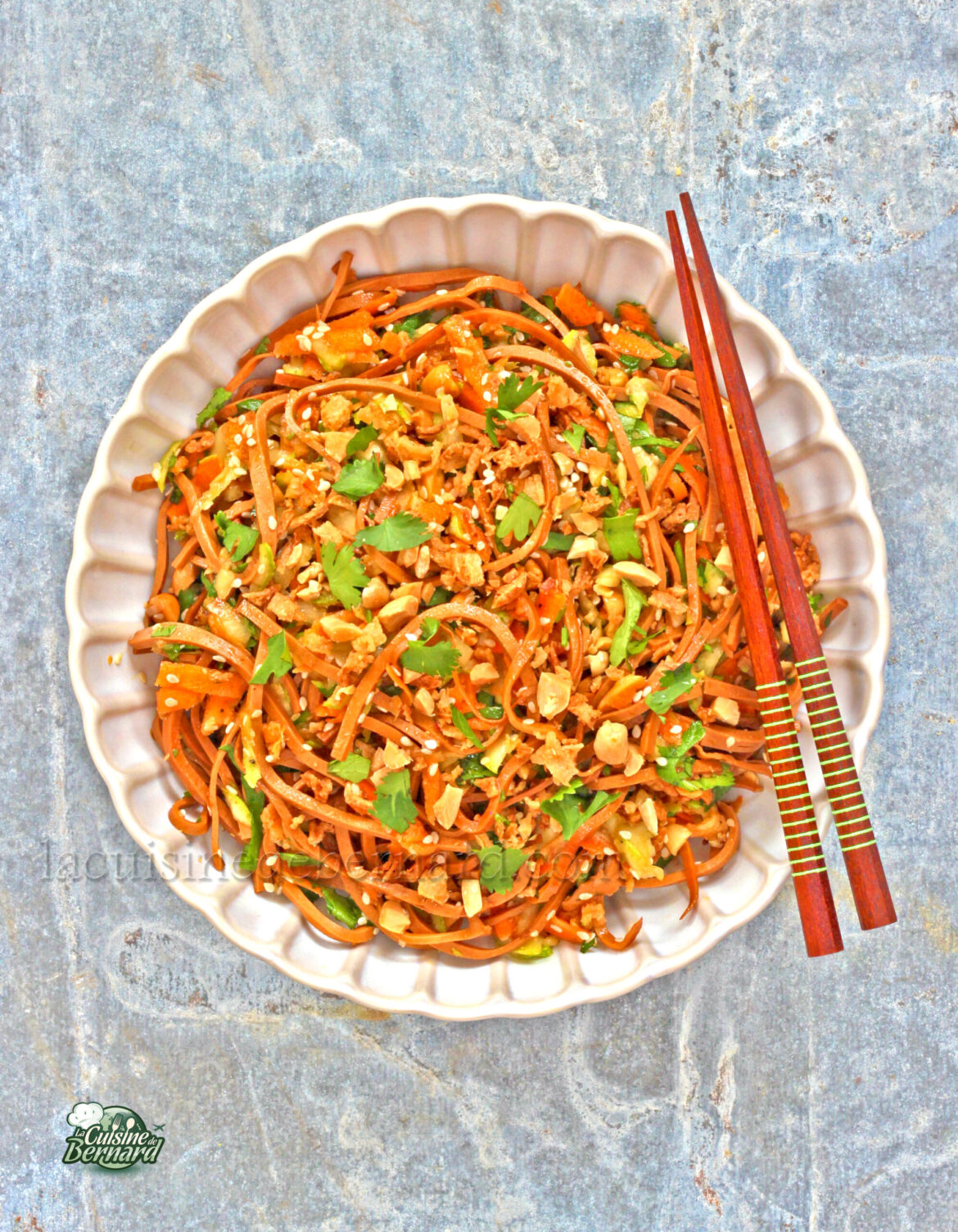 Un bol de nouilles sautées garnies d'arachides hachées et d'herbes, inspiré de la salade végétarienne, servi avec des baguettes en bois sur une surface grise texturée.