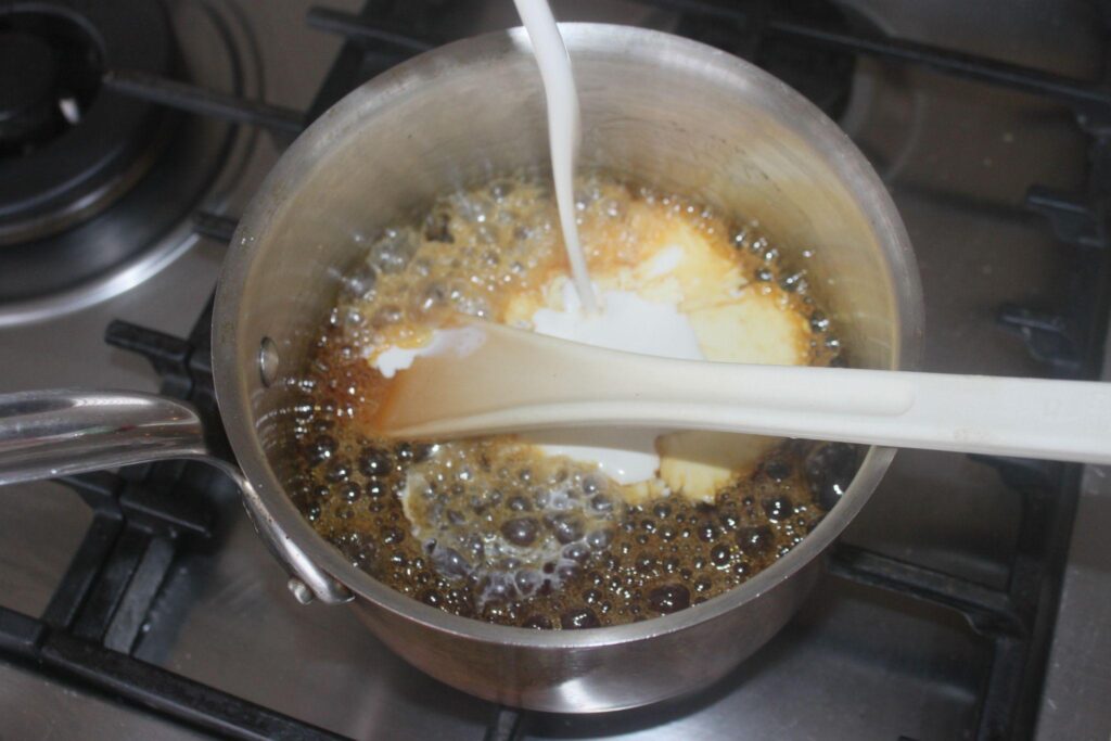 Une casserole de caramel bouillonnant sur une cuisinière avec de la crème versée et une spatule blanche mélangeant le contenu.