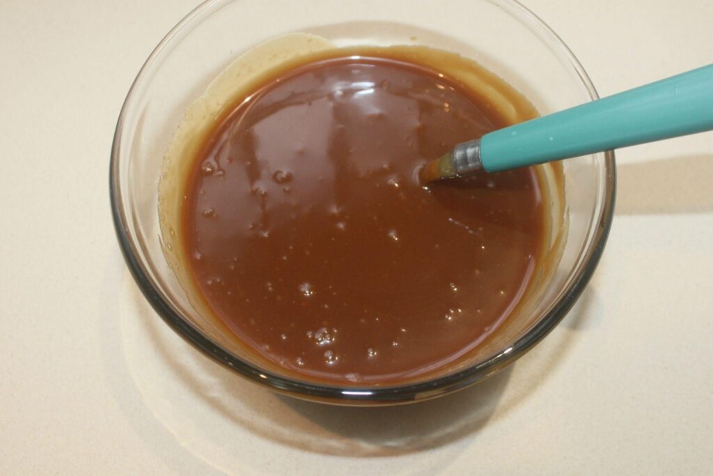 Un bol en verre rempli de sauce au caramel brun, avec une cuillère à manche bleu posée à l'intérieur.