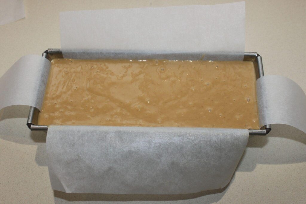 Un moule à pain rectangulaire recouvert de papier sulfurisé est rempli de pâte brun clair, prête à être cuite.